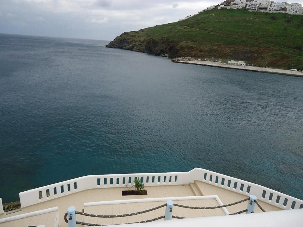 Vista costera desde un balcón con barandilla, sobre un mar tranquilo y una colina cubierta de hierba con edificios a lo lejos.
