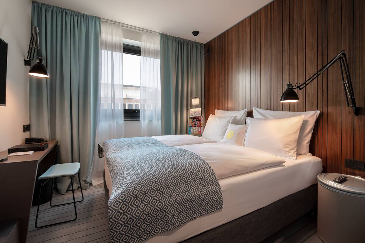 A modern hotel room with a large bed, a desk with a chair, a small trash bin, and a wall-mounted lamp.