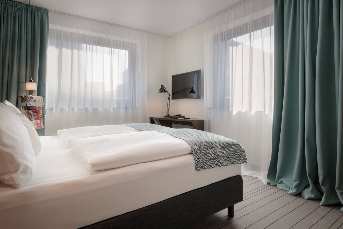 A modern hotel room with a large bed, a desk with a lamp, a flat-screen TV, and sheer curtains.