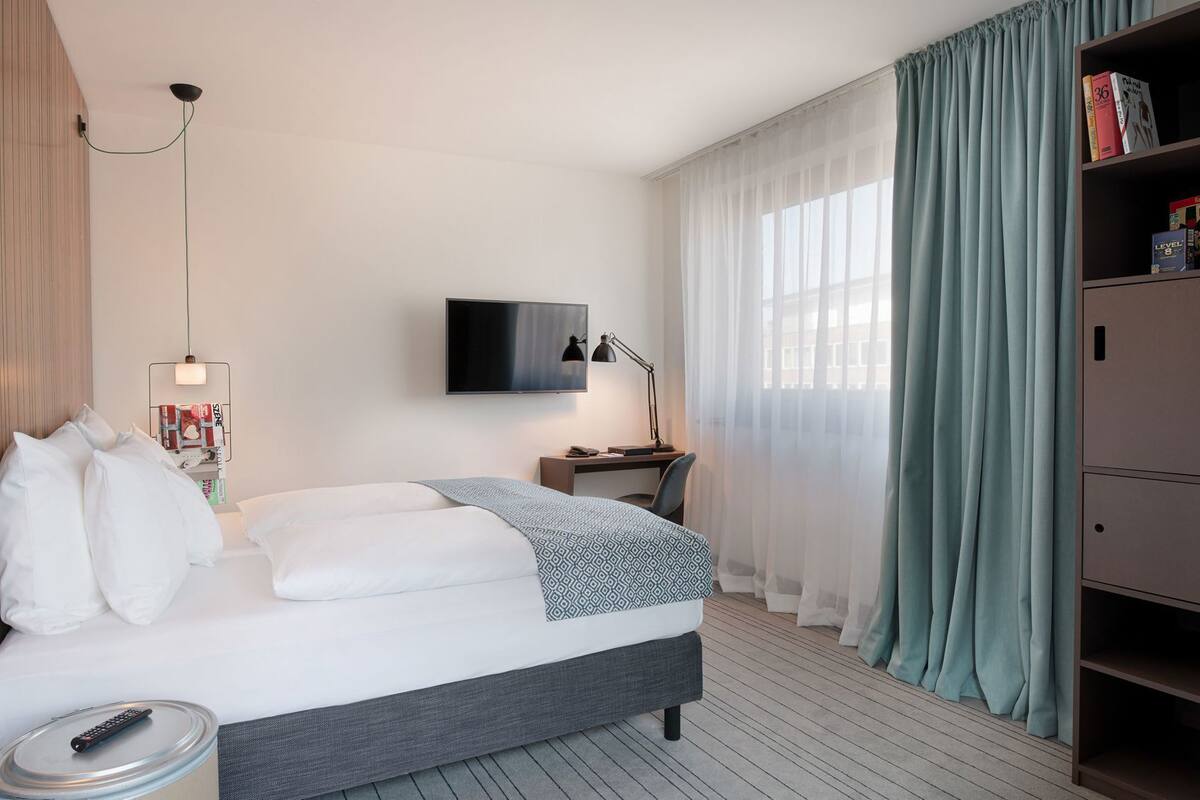 A hotel room with a bed, a desk, a TV, a bookshelf, and a window with curtains.
