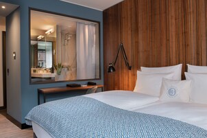 Ein modernes Hotelzimmer mit einem großen Bett, einer Wand mit Holzvertäfelung, einem Schreibtisch mit Stuhl, einem großen Spiegel und einer Nachttischlampe.
