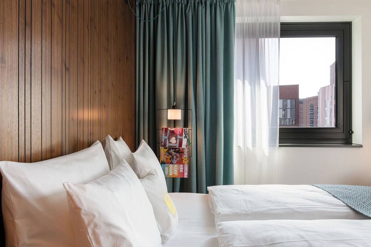 A hotel room with a wooden wall, a bed with white bedding, a window with a view of buildings, and a magazine rack with a magazine.