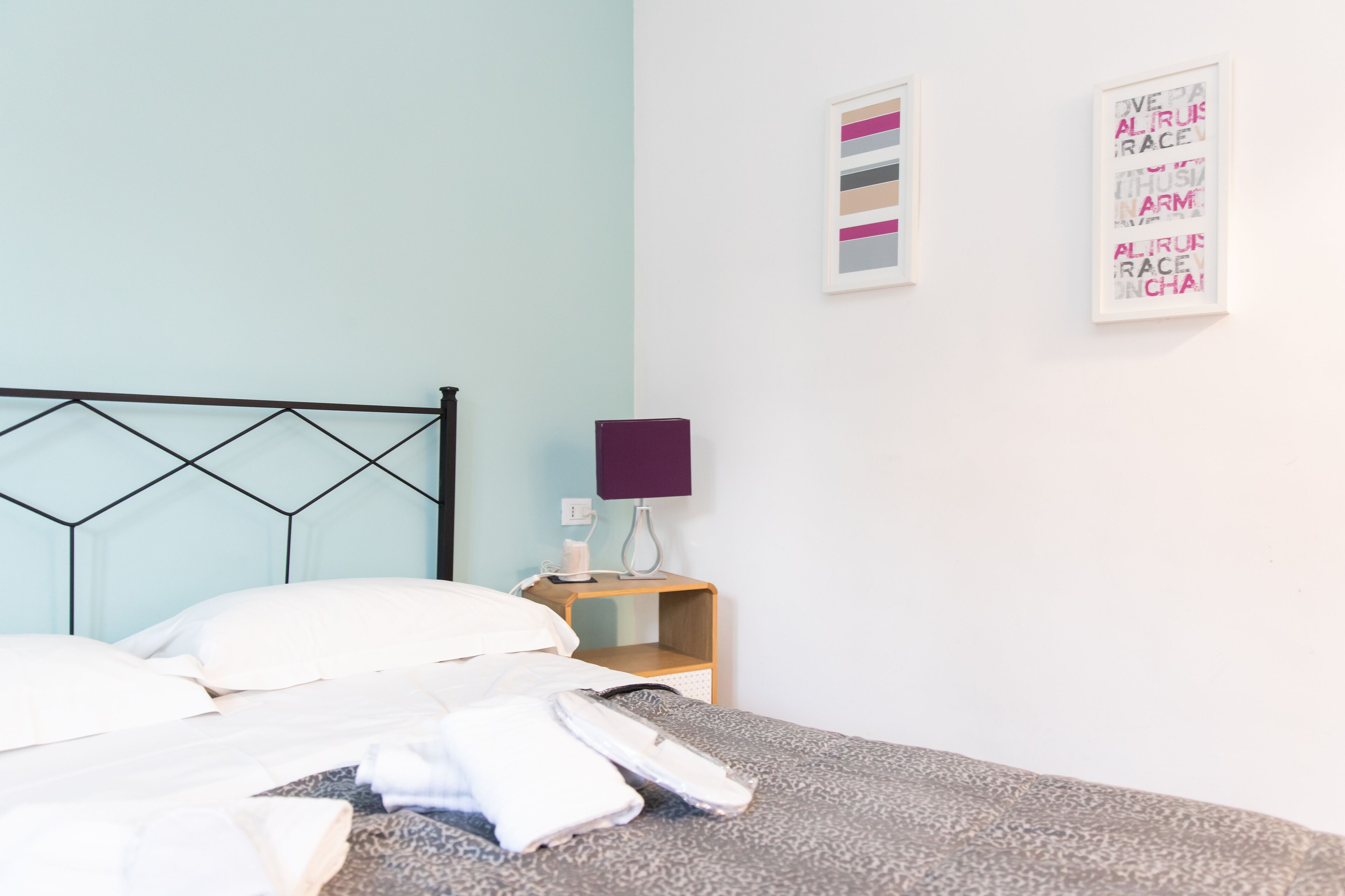 Een net opgemaakt bed met witte lakens, een houten nachtkastje met een paarse lamp, en twee ingekaderde kunstwerken aan de muur.