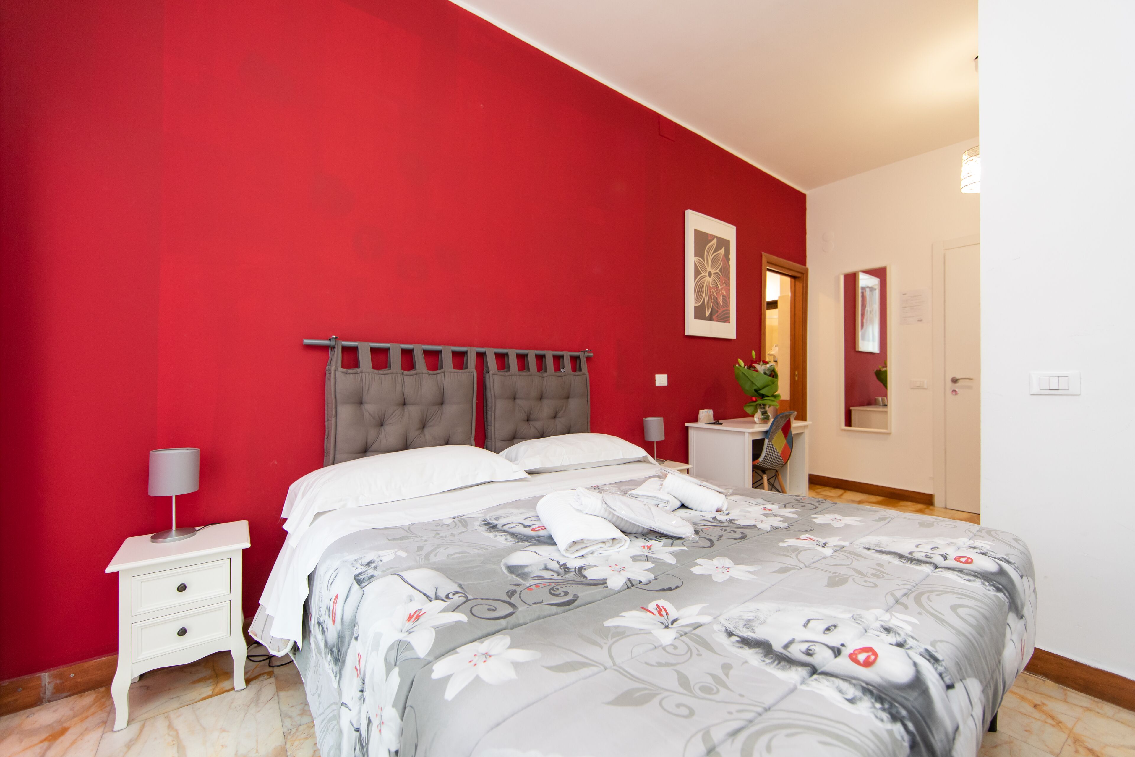 Une chambre avec un mur rouge, une tête de lit rembourrée de couleur grise, un couvre-lit blanc et une petite table de chevet avec une lampe.