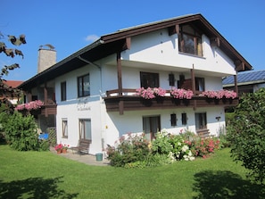 A two-story building with a balcony adorned with flowers, a sign indicating it is a guesthouse, and a well-maintained garden. - Gästehaus Am Buchenbichl (Schoenau am Koenigssee)
