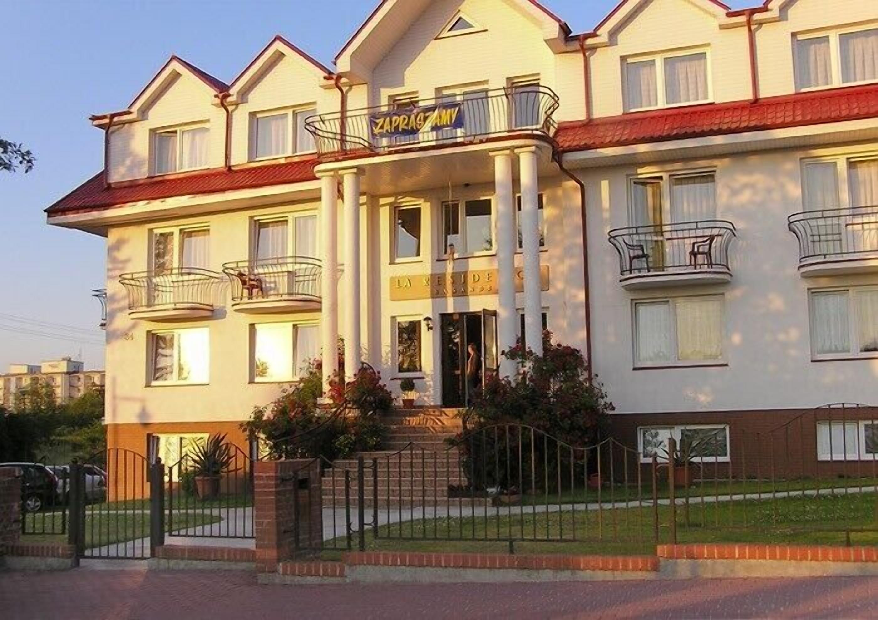 Een meerlaags hotel met een centrale ingang, balkons en een bord waarop staat dat het een hotel is.