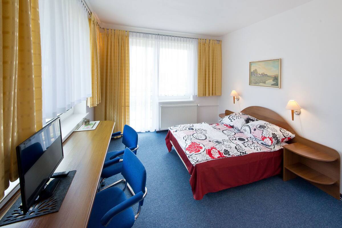 Een hotelkamer met een bureau, twee stoelen, een bed met een bloemenprint, een raam met gordijnen, een radiator en een wandlamp.