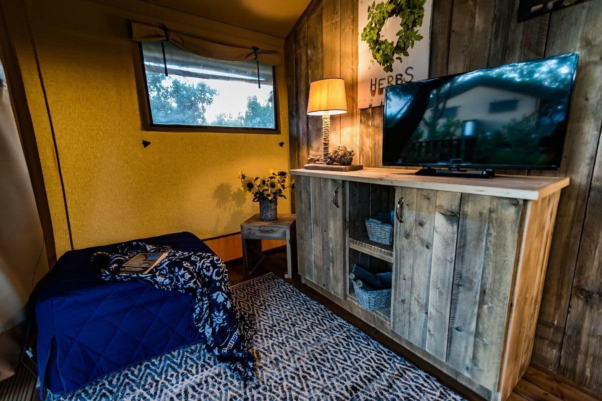 Ein gemütliches Zimmer mit einem Bett, einem Fernseher auf einer Holzkommode, einem kleinen Tisch mit einer Lampe und einem Fenster mit Blick auf Bäume.