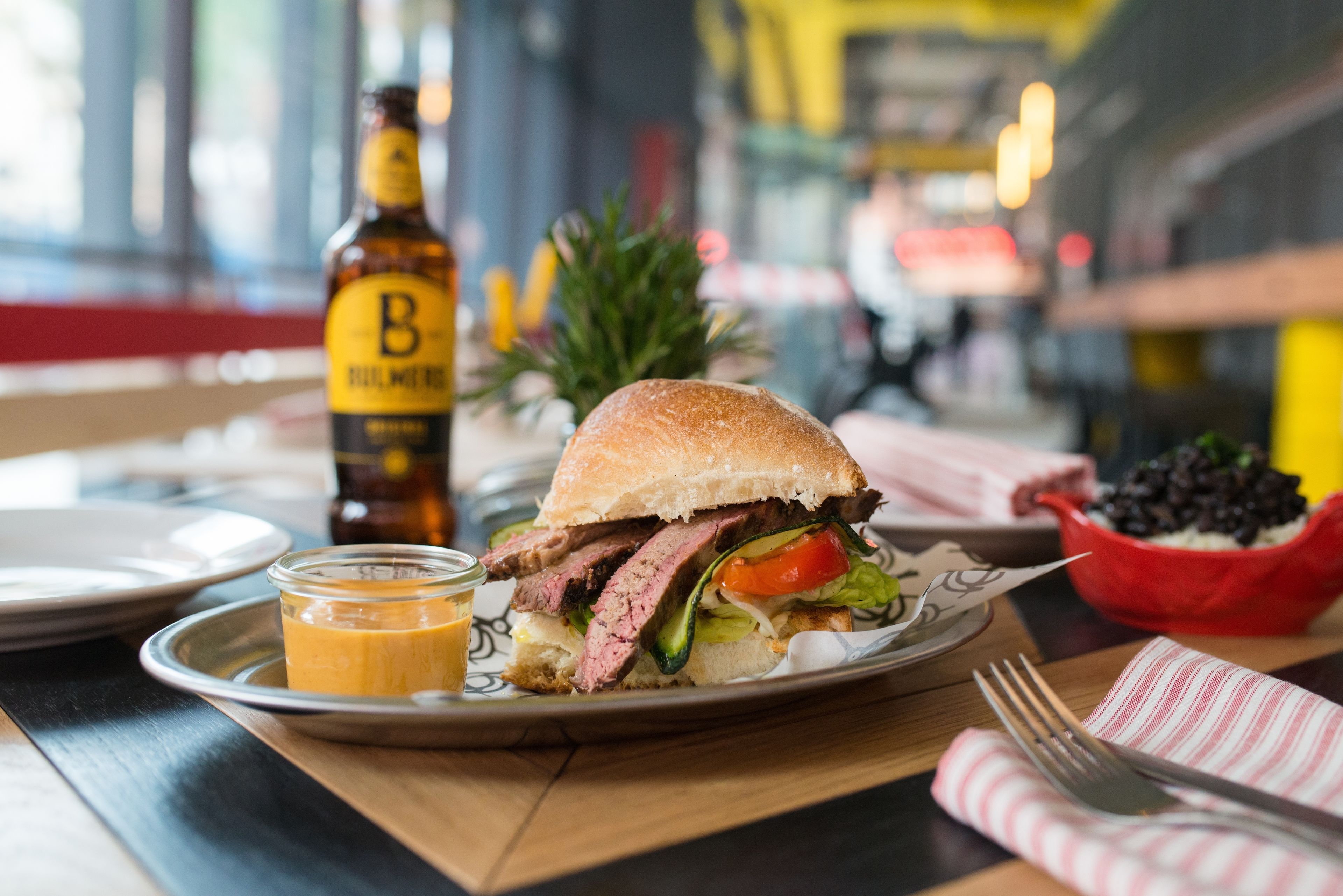 Un entrepà a un plat, amb una ampolla de cervesa i condiments al fons.