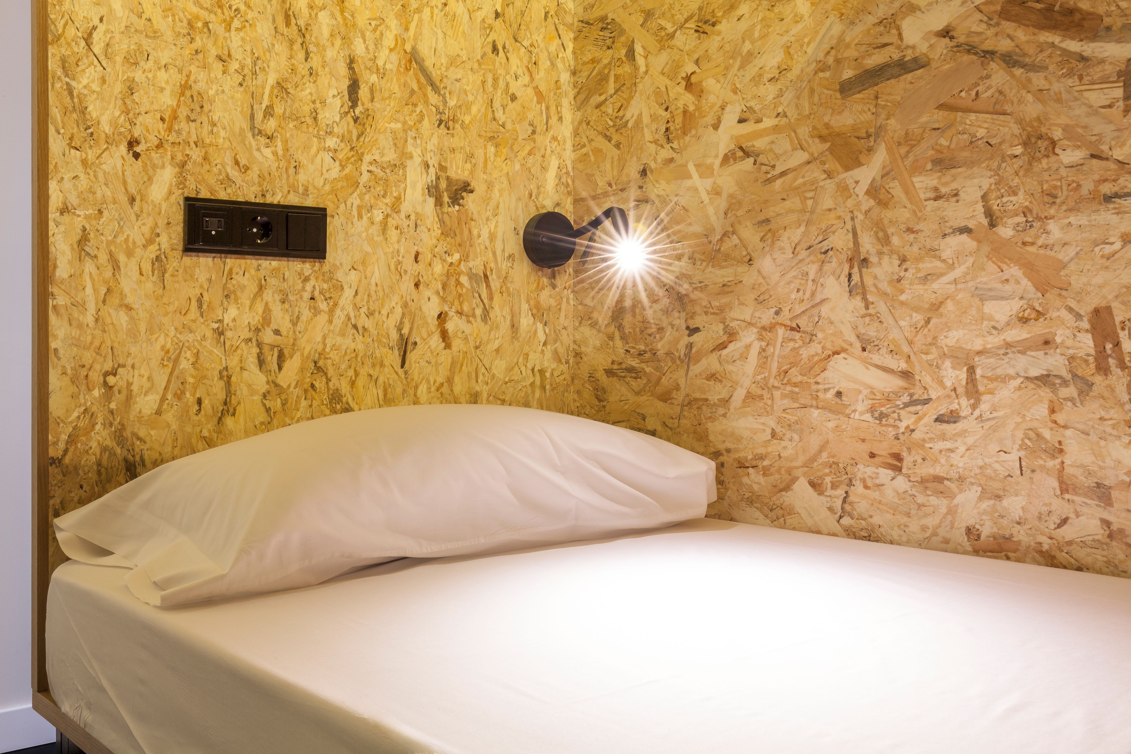 Una cama con colchón y almohada blancos, una lámpara de pared y un enchufe en una pared con textura.