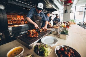A commercial kitchen with chefs preparing food, a meat roasting station, and various food items and condiments on the counter.