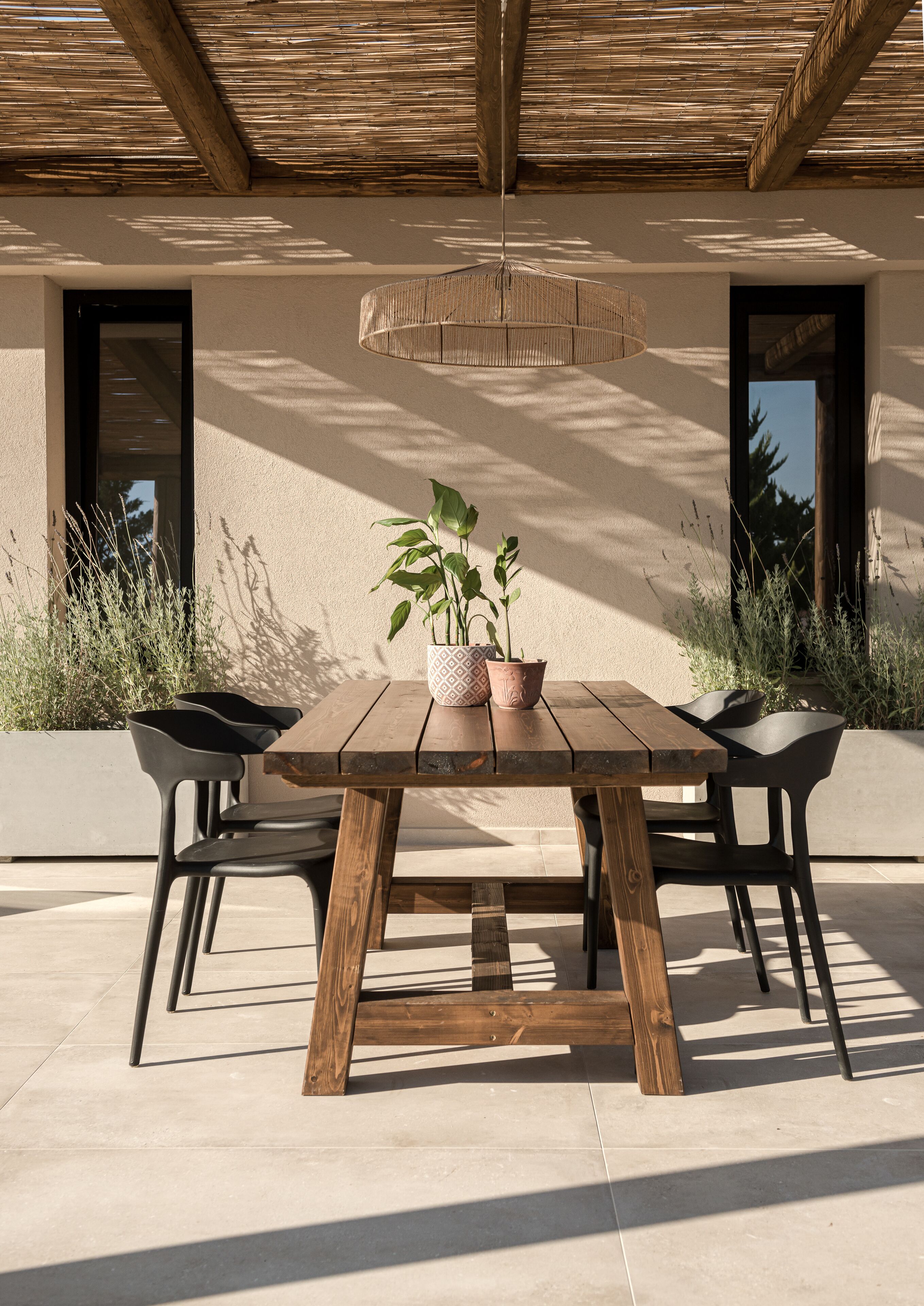 Pátio externo com mesa e cadeiras de madeira, rodeado por jardineiras e uma estrutura de pérgola.