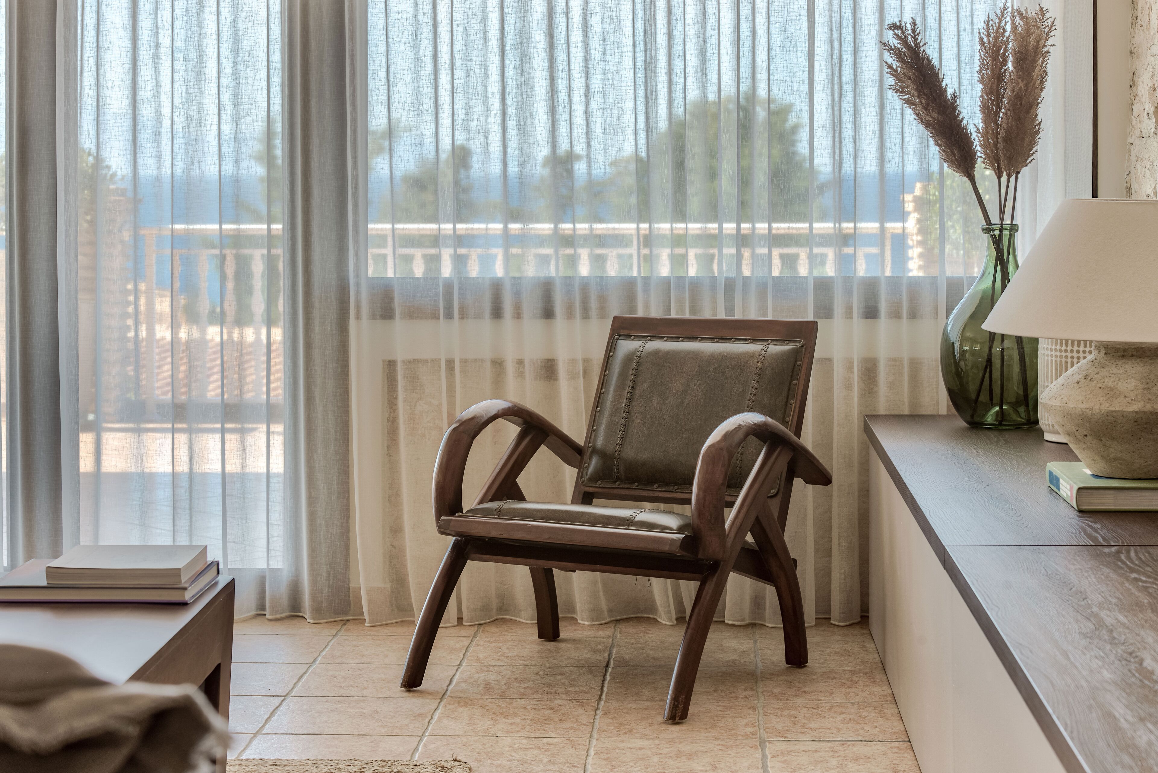 Una habitación con vista al mar, una silla de madera, una mesa con libros y un jarrón con plumas de miscanthus.
