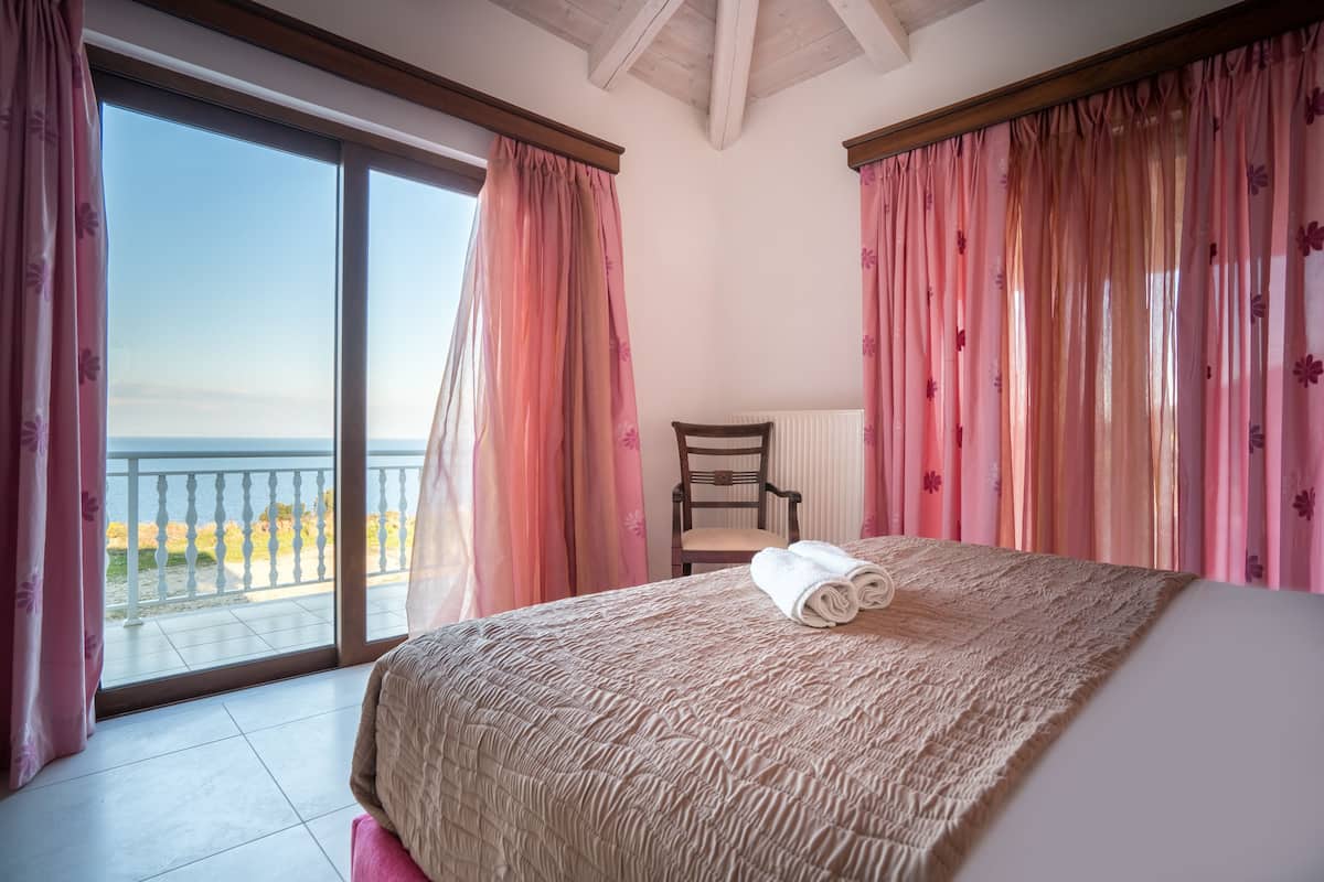 A bedroom with a large bed, a chair, and a view of the sea through a sliding door.