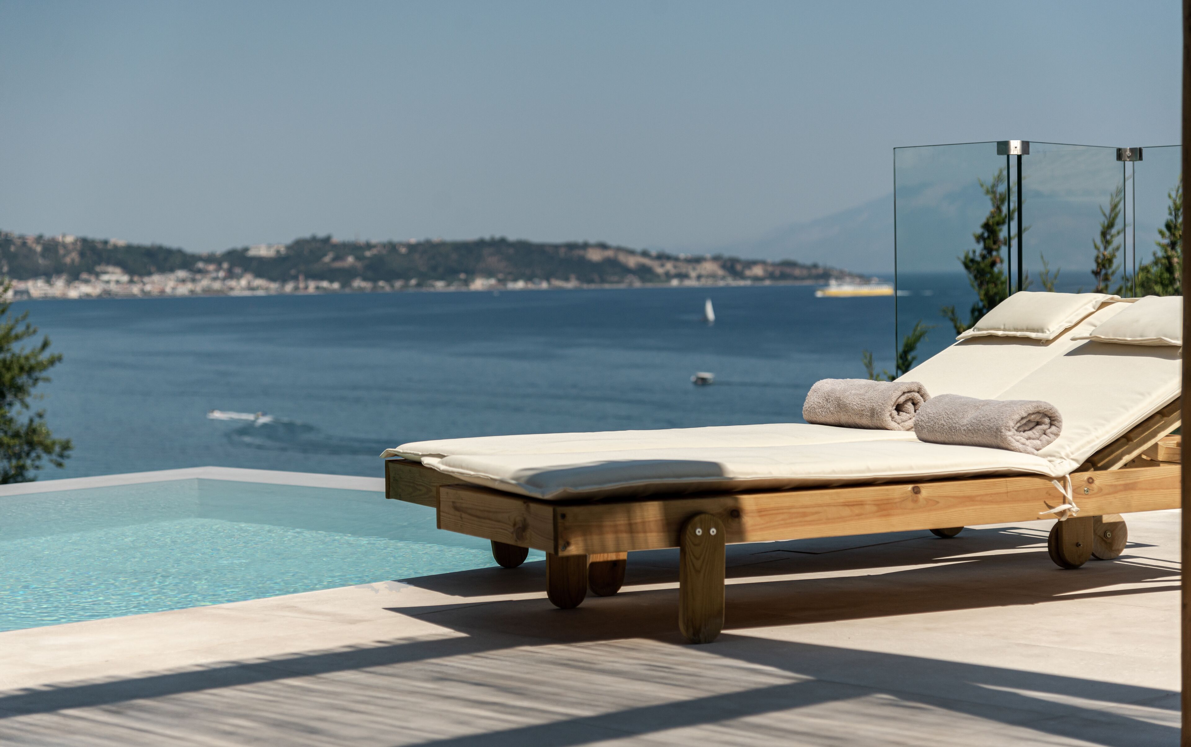 Una silla de lounge de madera con un cojín blanco y toallas enrolladas, con vista a una piscina y al mar.