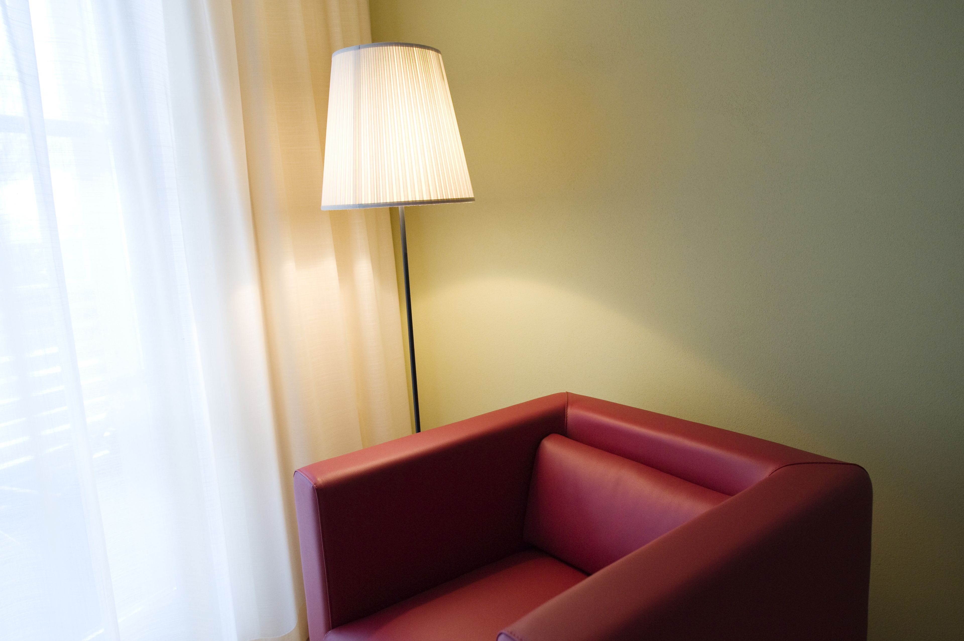 Une chaise rouge et un lampadaire dans une pièce dont le mur est de couleur claire et qui possède une fenêtre ornée de rideaux légers.