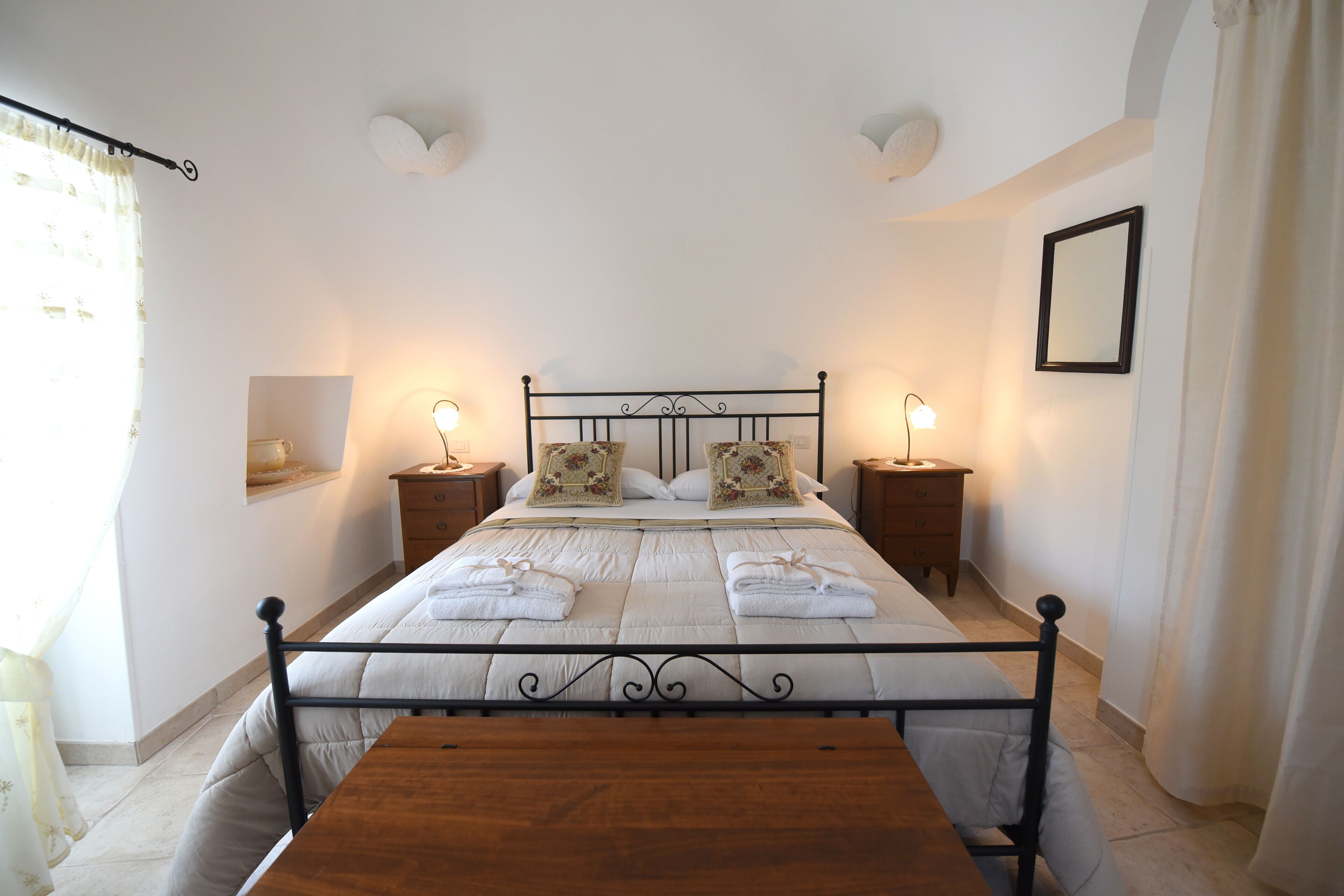 A neatly made bed with pillows and towels, two bedside tables with lamps, a wooden bench, and a wall-mounted mirror.
