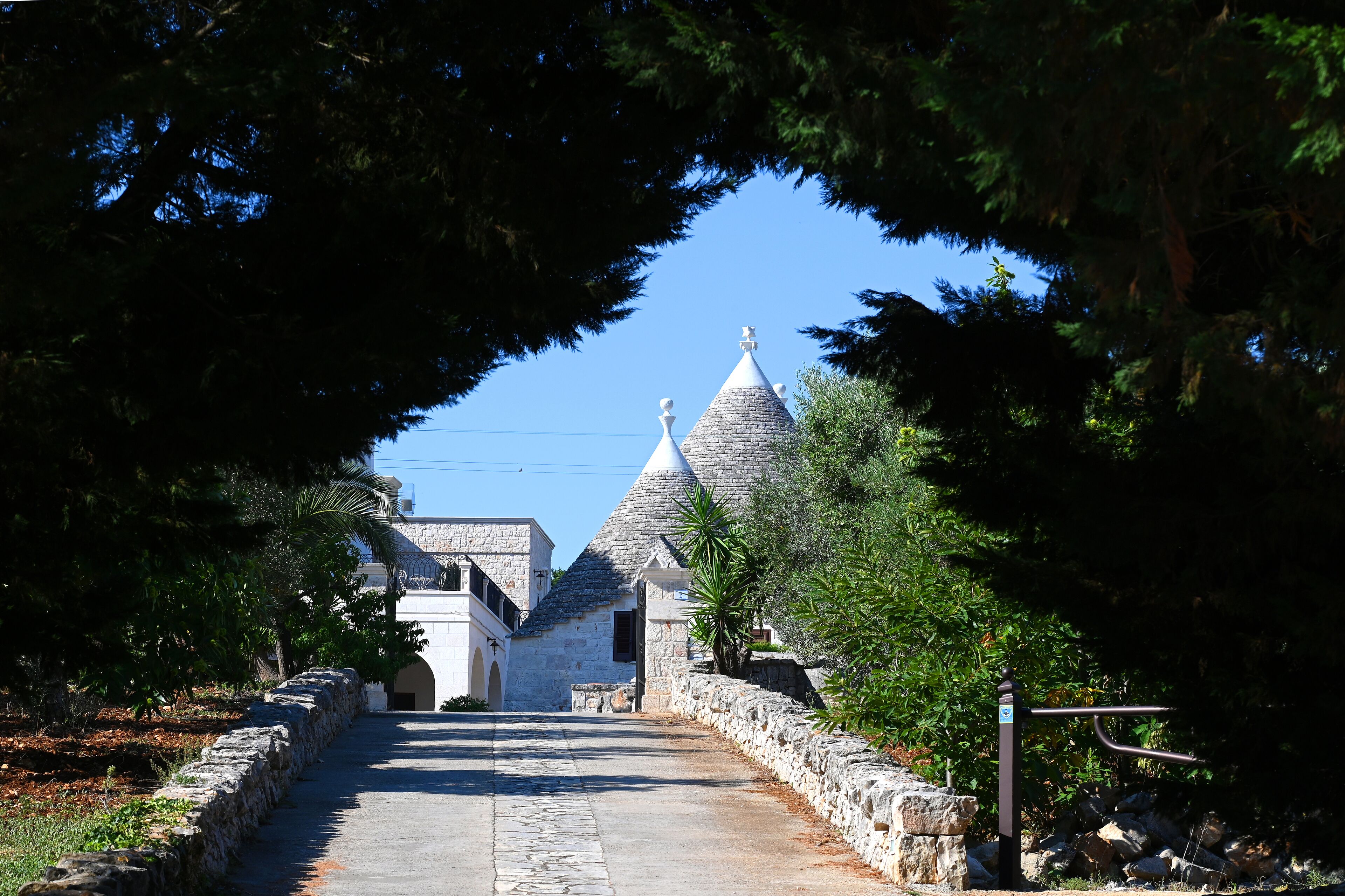 A stone pathway leading to a building with a conical roof, surrounded by trees and a stone wall.