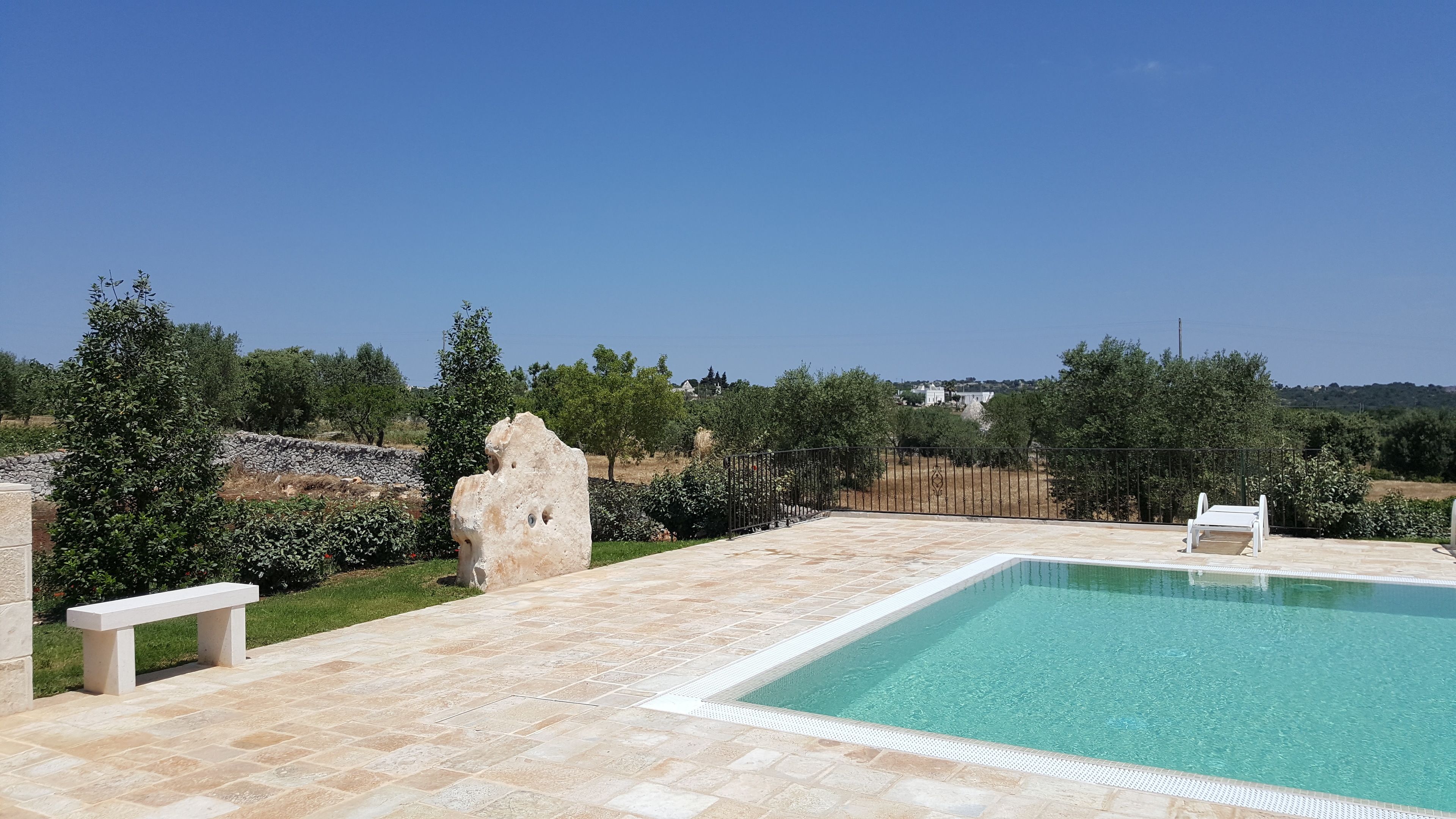 A swimming pool with a stone deck, surrounded by greenery and a stone wall.