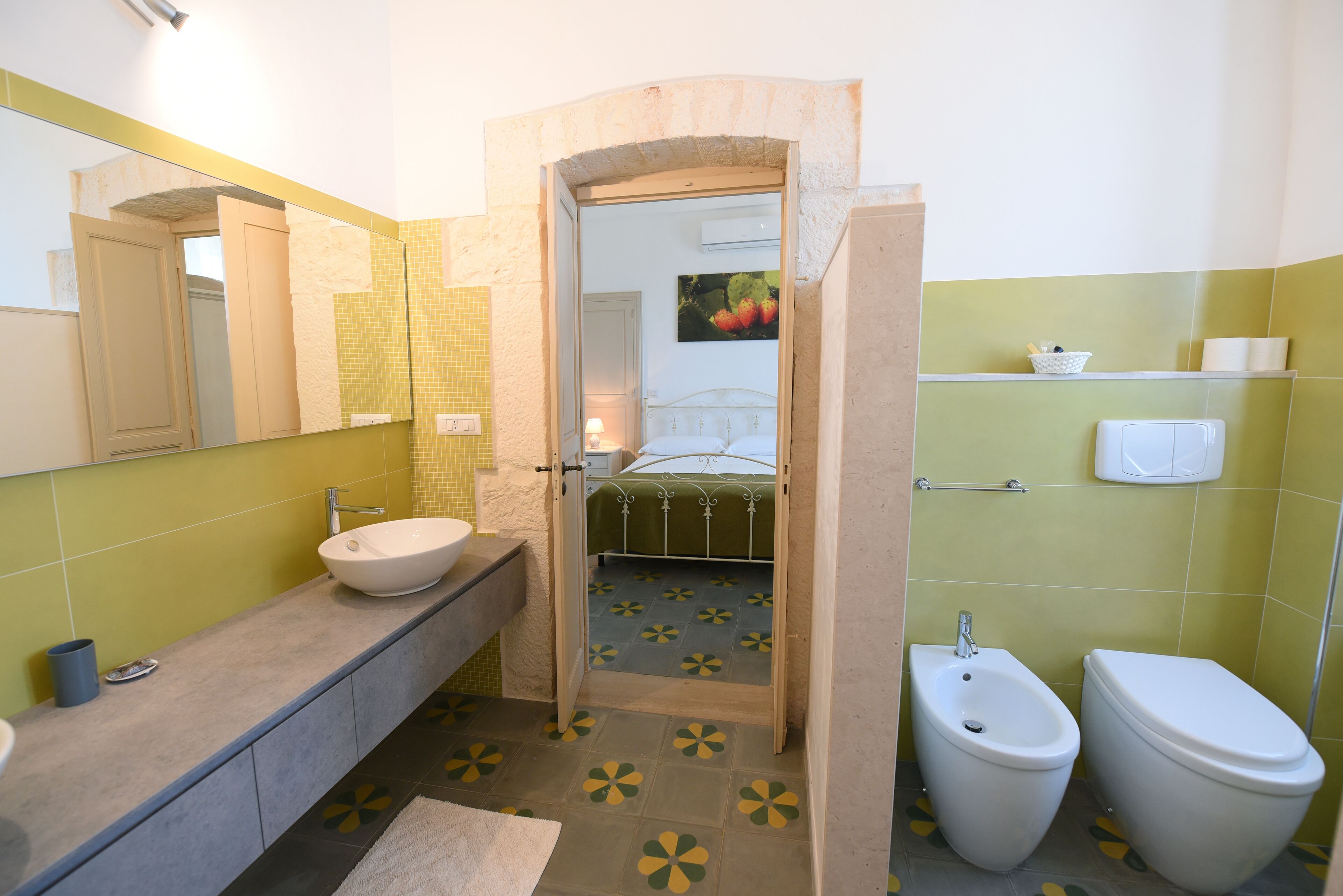 Baño con lavamanos, bidé e inodoro. Hay un espejo sobre el lavamanos y una puerta que da a un dormitorio.