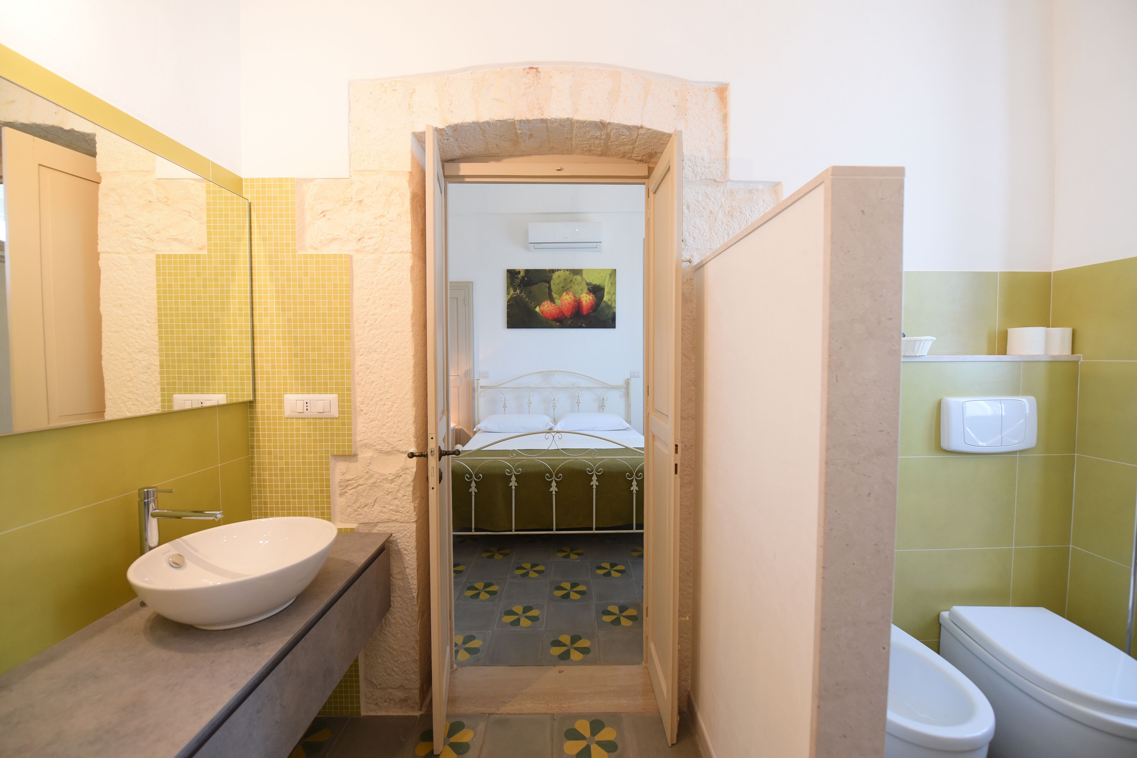 Ein Badezimmer mit Waschbecken, Toilette und einem Bett, das durch eine offene Tür zu sehen ist.