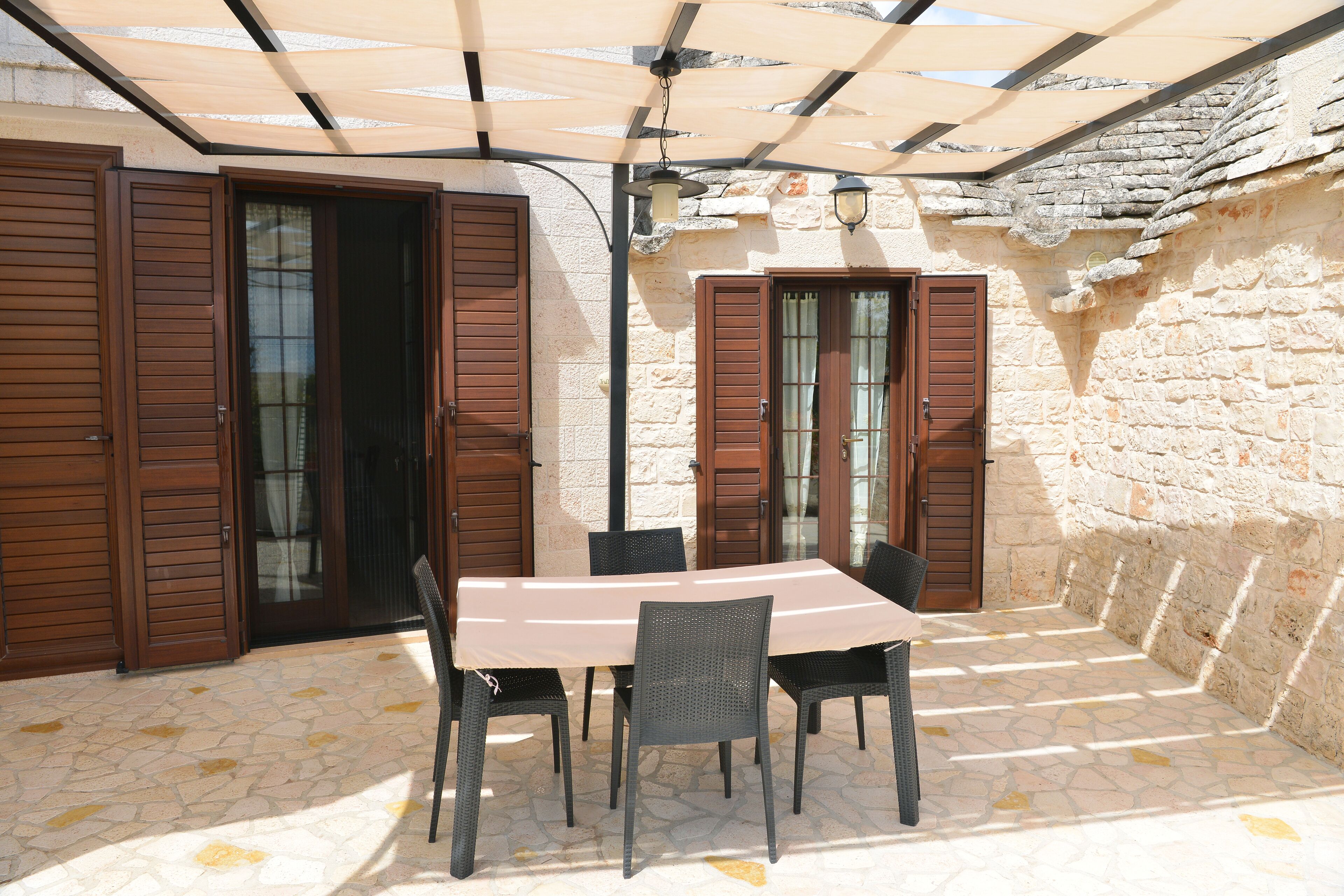 Une terrasse extérieure avec une table et des chaises, un sol en pierre, des volets en bois et un mur en pierre.