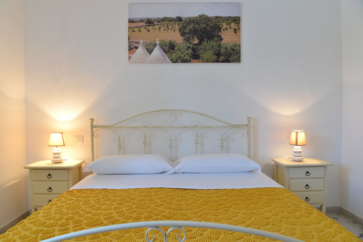 A bed with a yellow patterned bedspread, two bedside lamps, and a wall-mounted picture of a landscape.