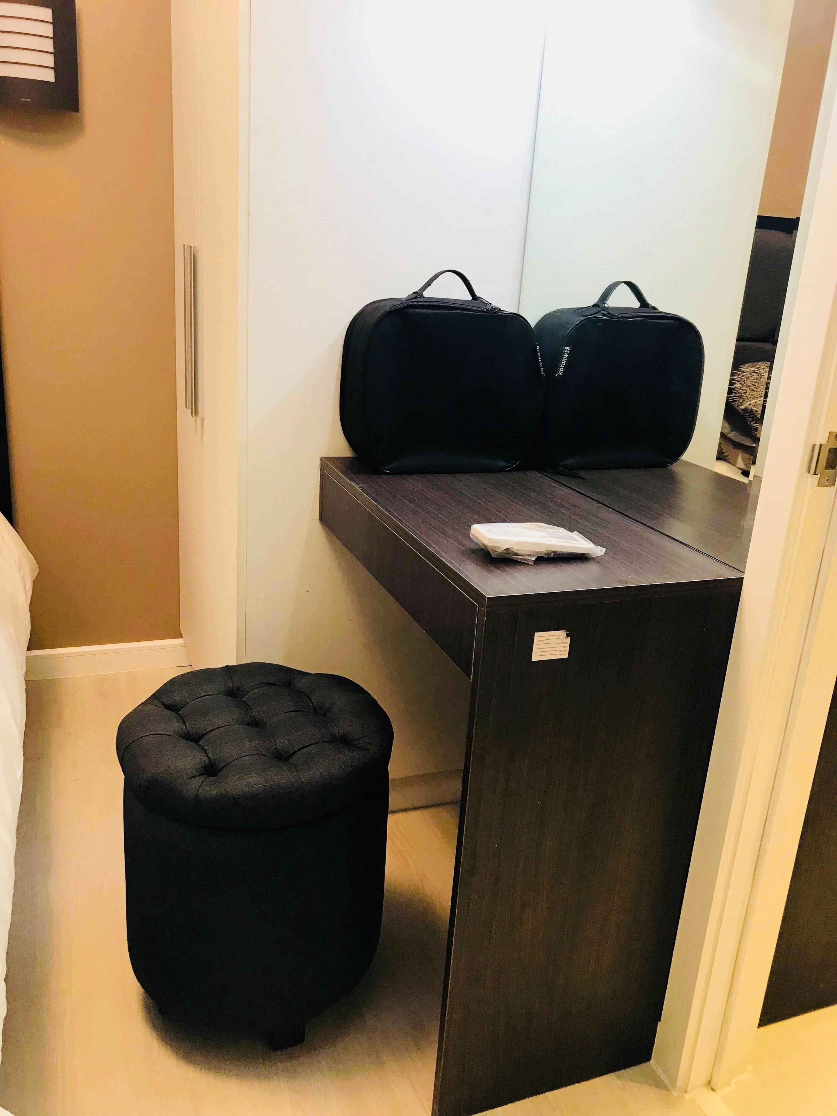 Une pièce de taille modeste, avec un bureau en bois foncé, deux sacs de sport noirs posés dessus, un petit pouf noir et un objet blanc sur le bureau.