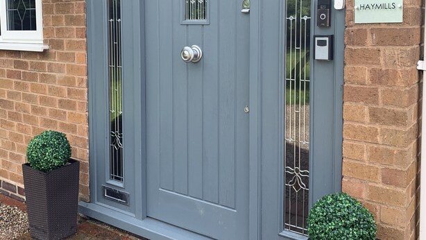 A grey front door with a small window and a doorbell. There are two potted plants on either side of the door. A sign on the wall reads "Haymills". - Haymills Guesthouse (Solihull)