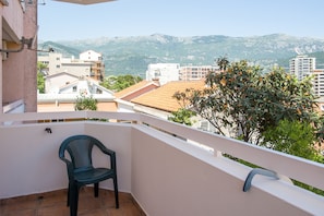 A balcony with a chair, a white railing, and a view of buildings and mountains in the background. - City Apartments (Budva)