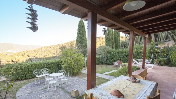An outdoor patio with a table and chairs, surrounded by greenery and a view of hills.