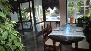 A dining area with a table set for two, chairs with cushioned seats, and a view of the street through large windows. - Hotel Freskia (Sarandë)