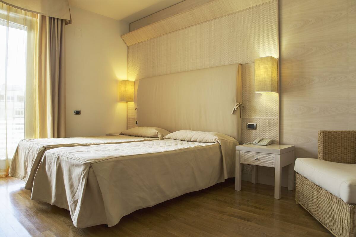 A hotel room with two beds, a bedside table with a lamp, a chair, and a large mirror.