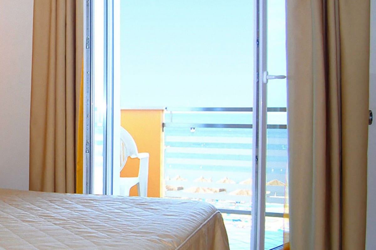 A hotel room with a bed, a nightstand, a lamp, and a view of the beach through the window.
