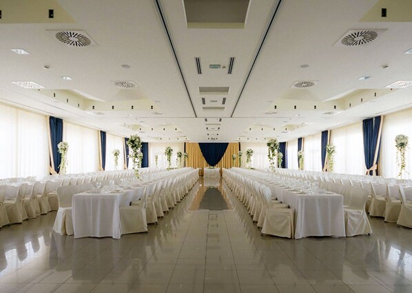 Une grande salle, bien éclairée, avec des rangées de tables et de chaises, une allée centrale et des compositions florales décoratives.