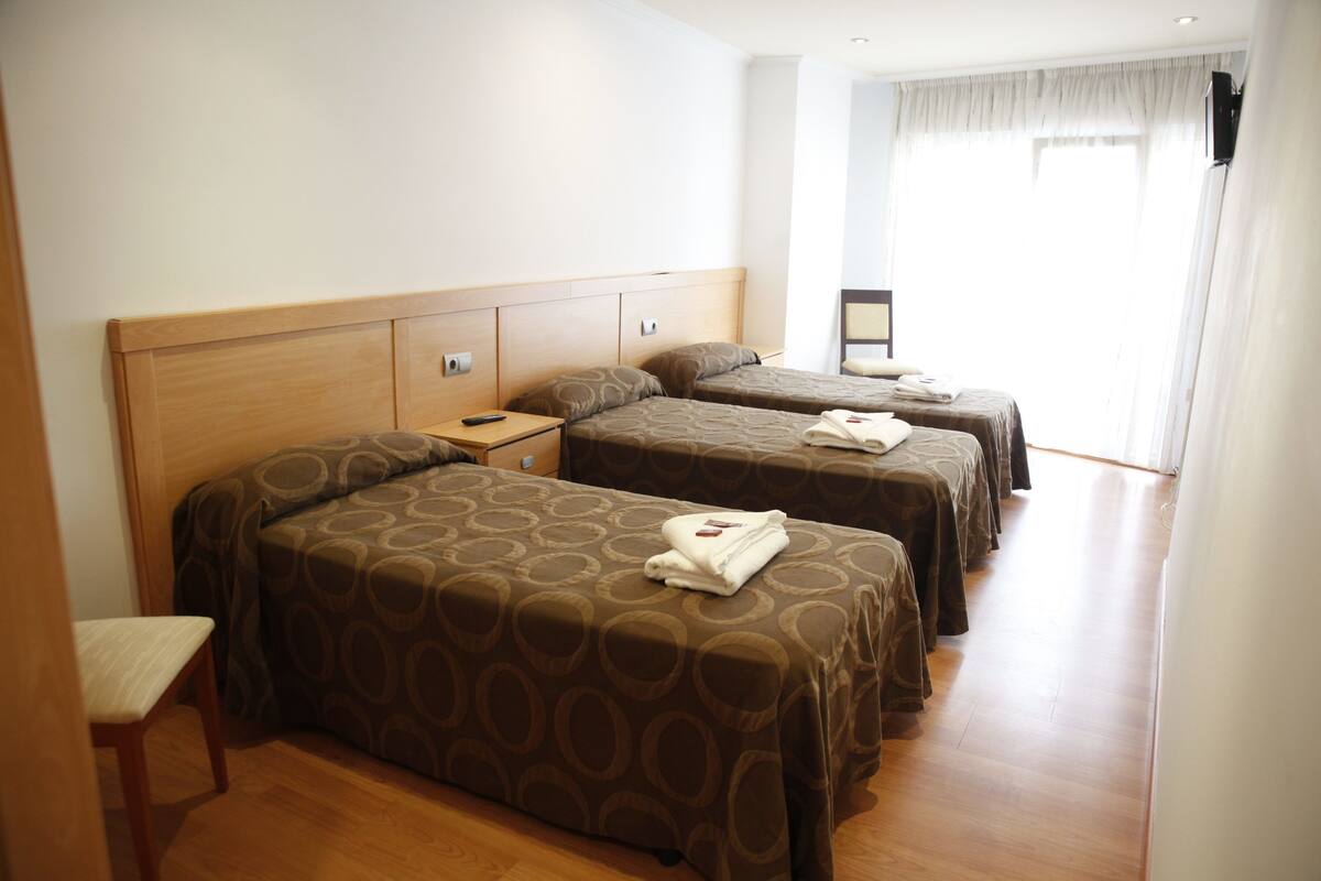 Una habitación de hotel con dos camas individuales, un cabecero de madera, una mesita de noche pequeña con un control remoto y una ventana con cortinas.