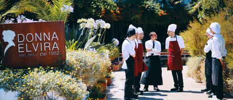 A group of chefs standing in front of a sign for Donna Elvira Ristorante dal 1952, surrounded by potted plants and flowers.