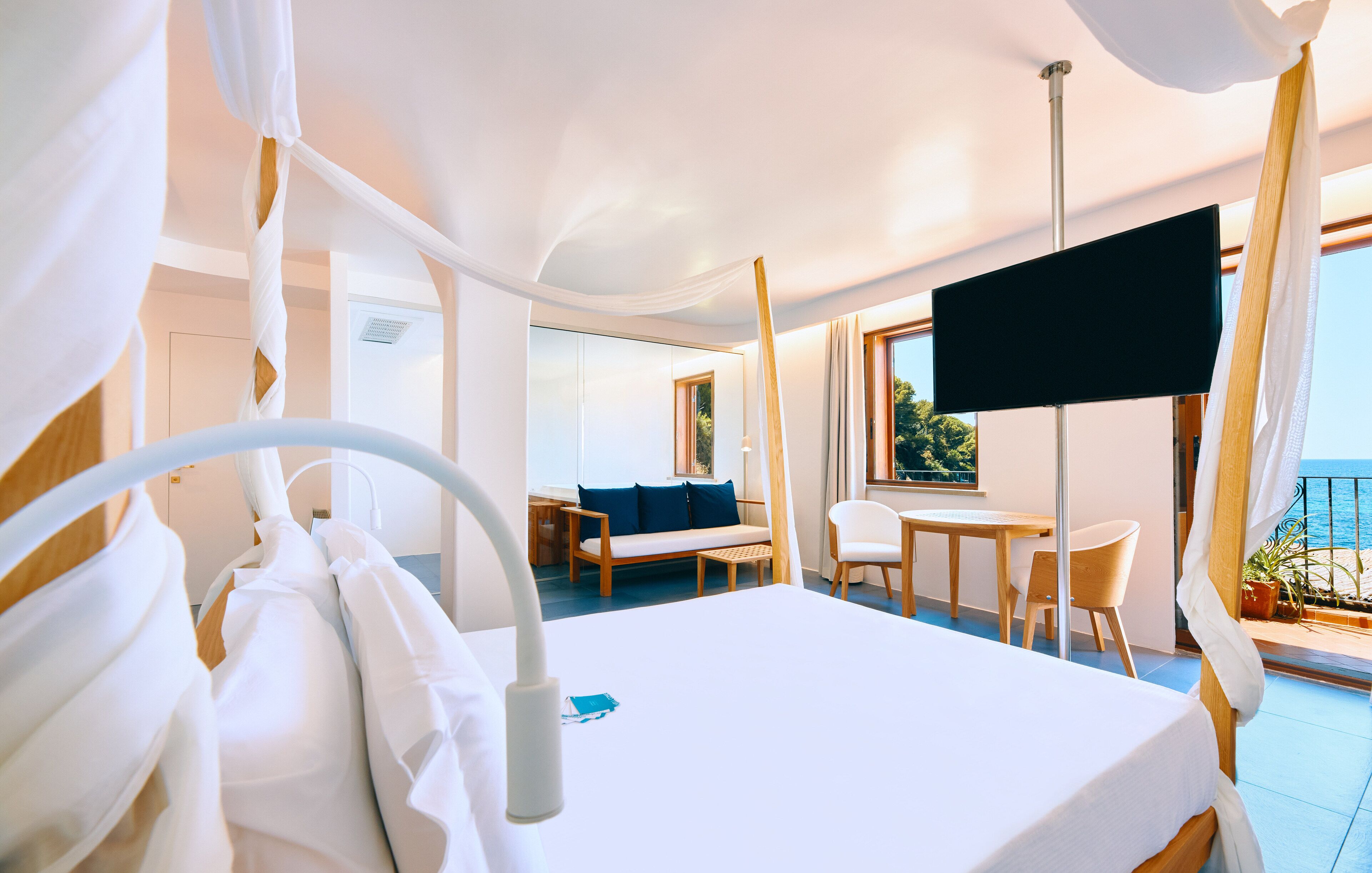 A hotel room with a bed, a desk, a chair, a sofa, a TV, and a balcony with a view of the sea.