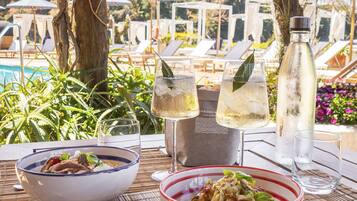 A dining table with two bowls of food, a wine glass, and a water bottle, set against a backdrop of a pool and lounge chairs.