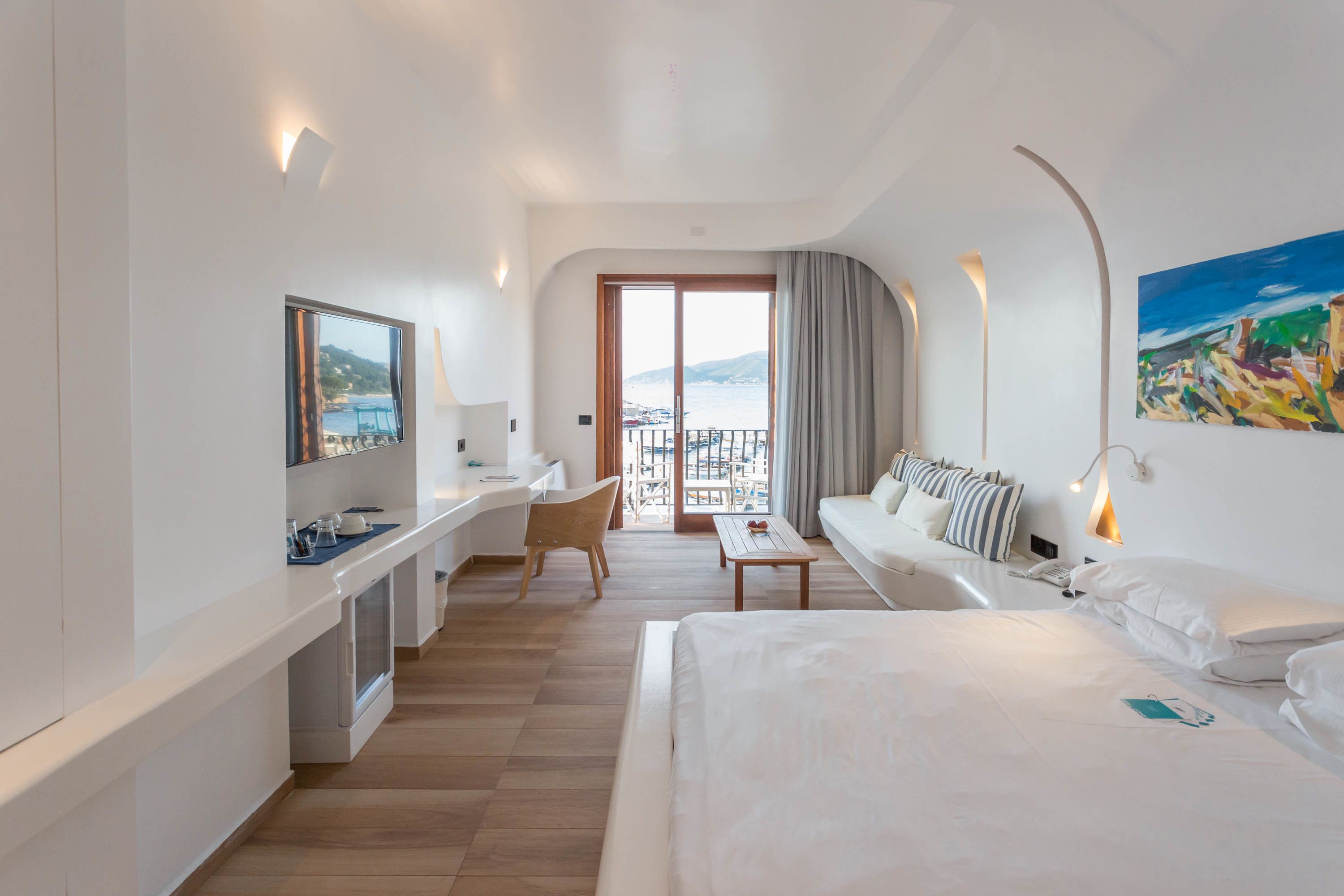 A modern hotel room with a large bed, a desk, a chair, a TV, and a balcony with a view of the sea.