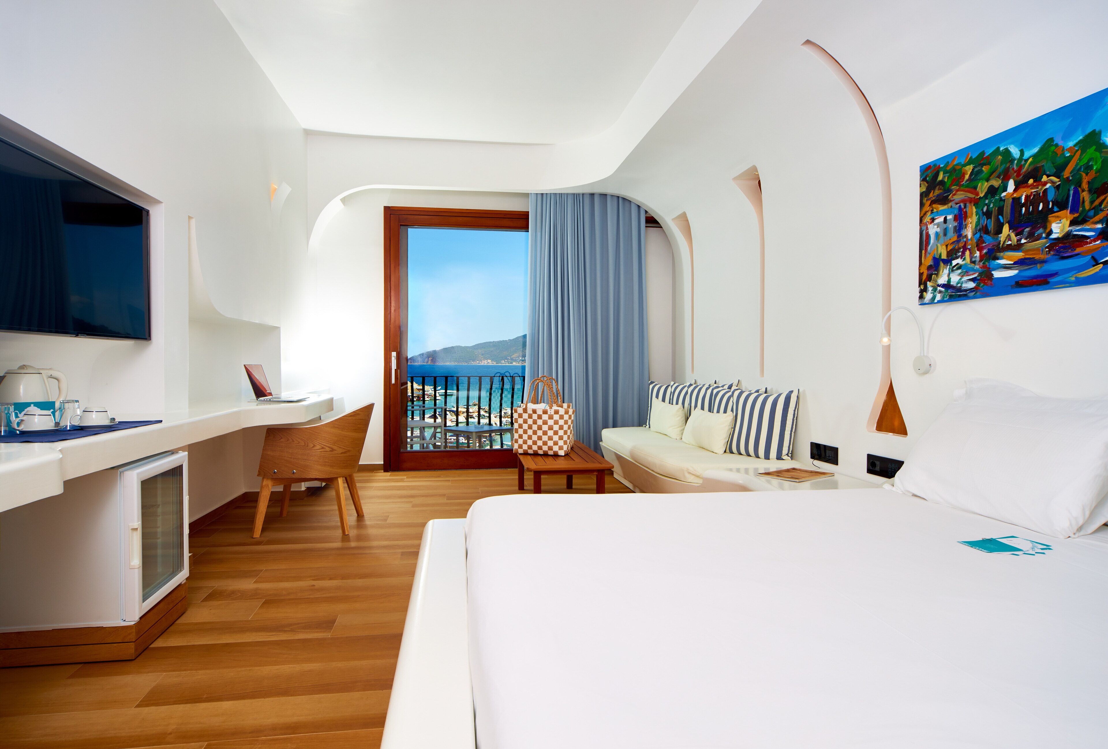 A modern hotel room with a bed, a desk, a chair, a small table, and a balcony with a view of the sea.