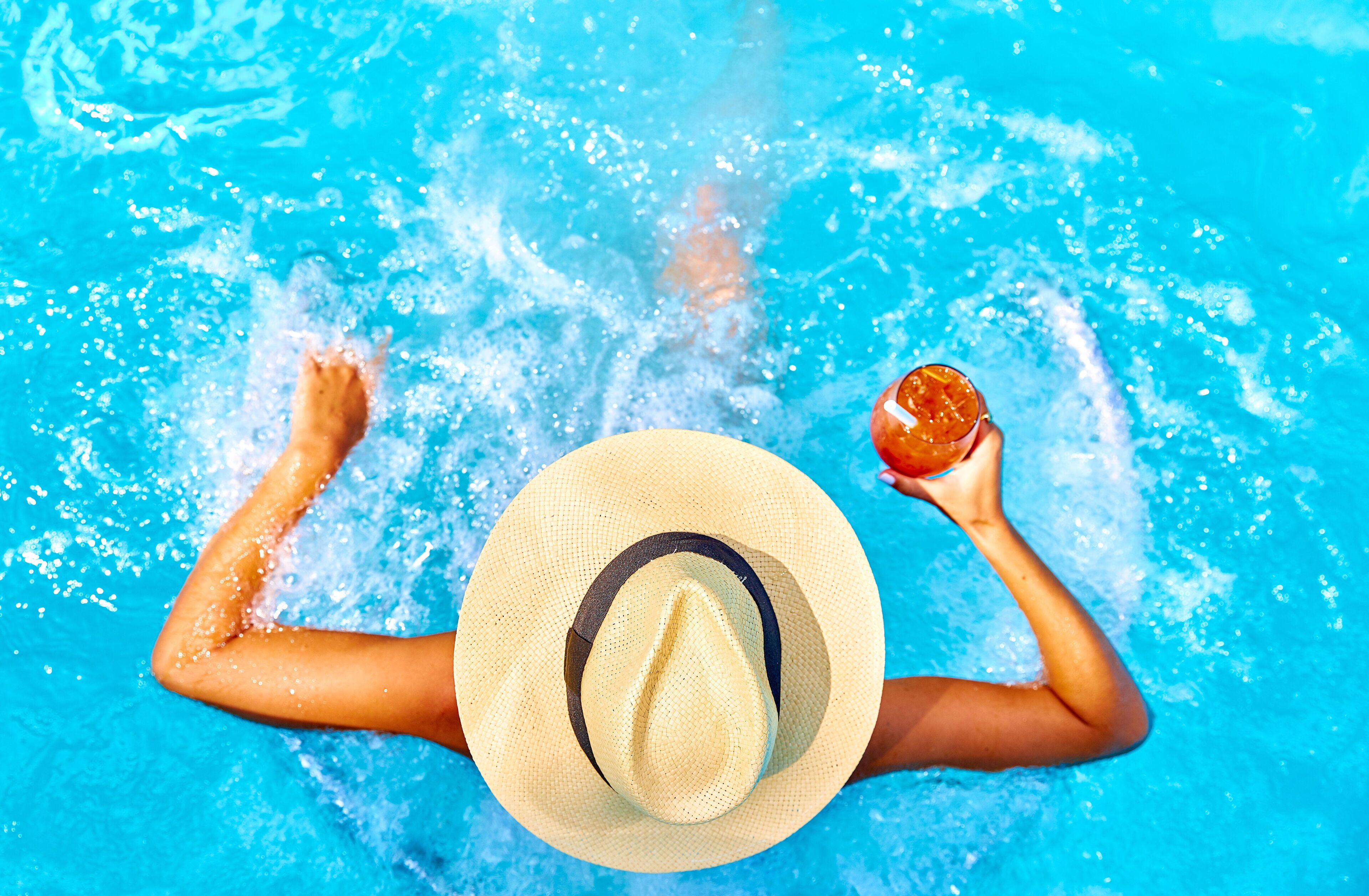 Una persona en una piscina, con sombrero de paja y sosteniendo una bebida.