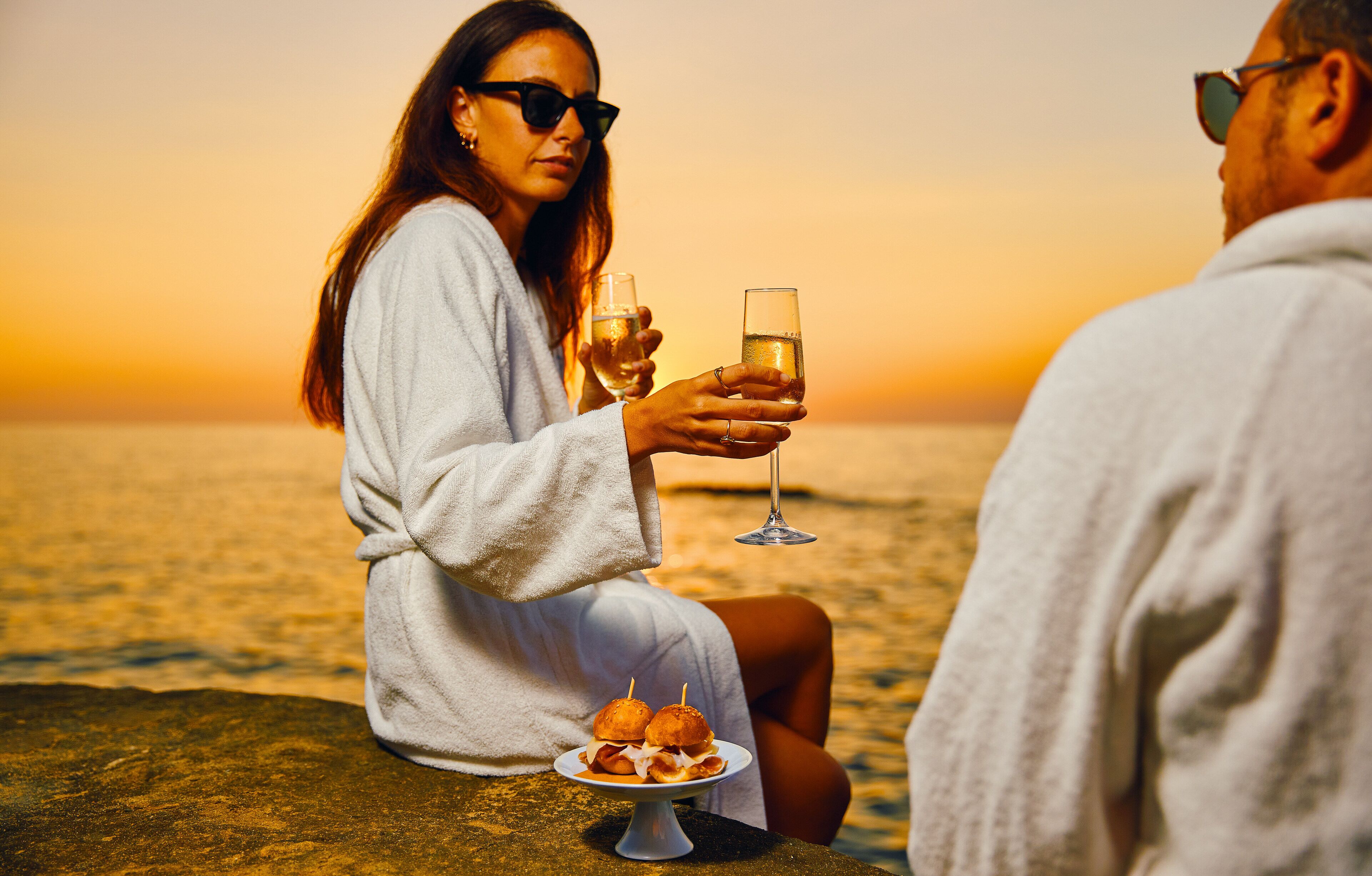 Two people sitting on a rock by the sea, holding glasses of champagne, with a plate of food in front of them.
