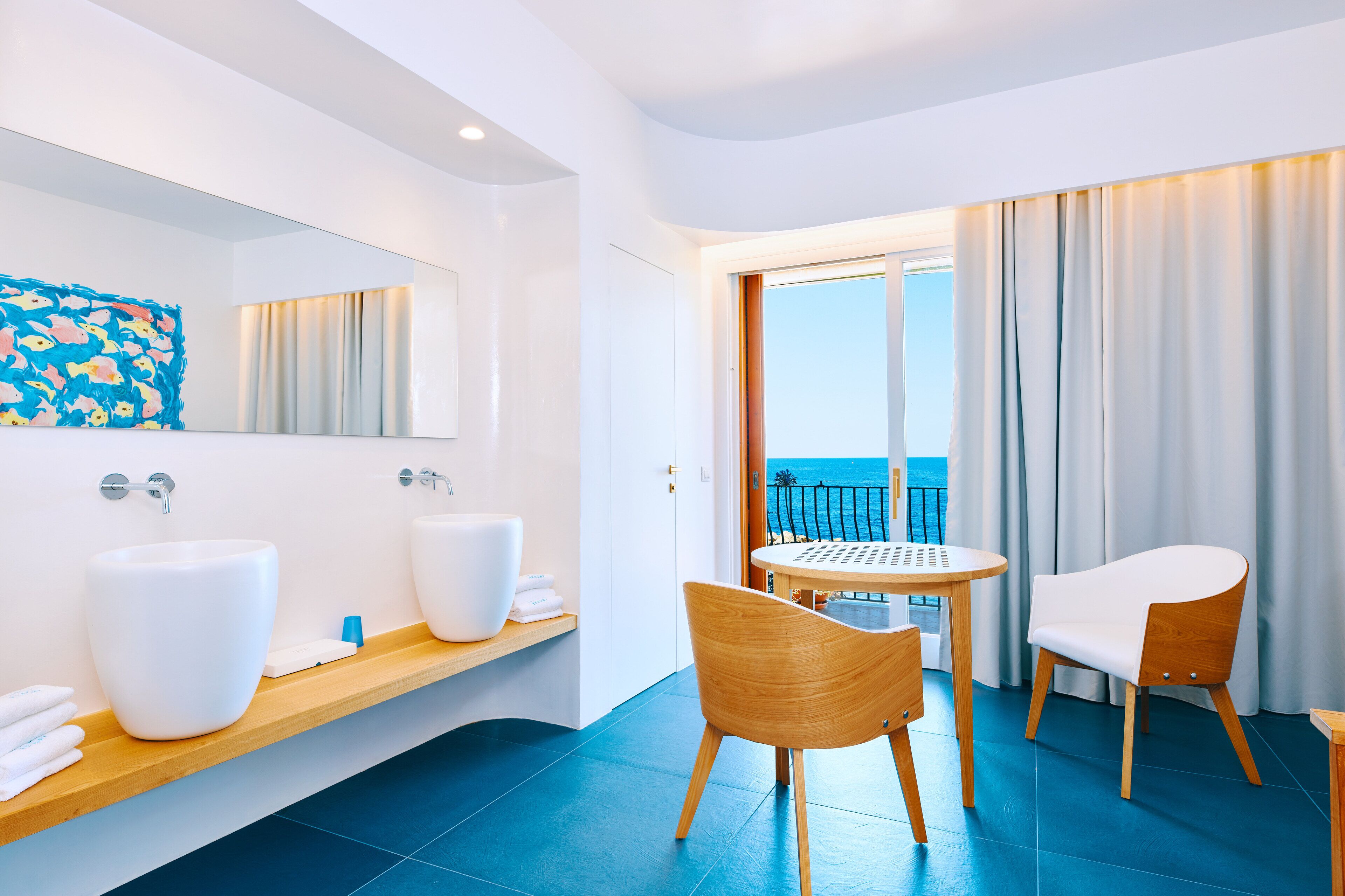 A modern bathroom with two large white sinks, a wooden vanity, and a large mirror. There is a wooden chair and a small round table with a view of the ocean through a glass door.