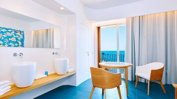 A modern bathroom with two large white sinks, a wooden vanity, and a large mirror. There is a wooden chair and a small round table with a view of the ocean through a glass door.