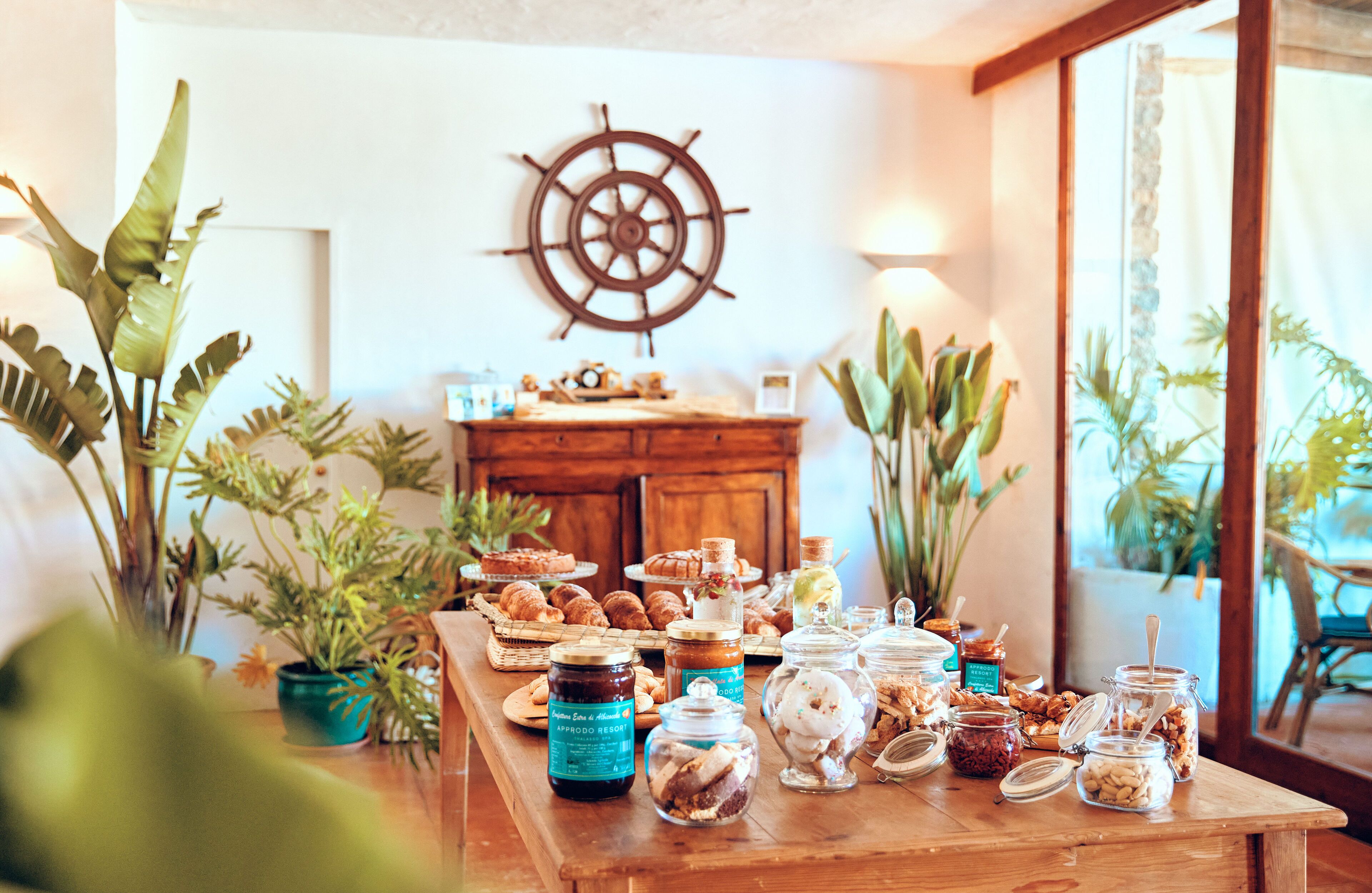 Un bufé de desayuno con diversos pasteles, mermeladas y untables sobre una mesa de madera, rodeado de plantas en macetas y una decoración de timón de barco en la pared.