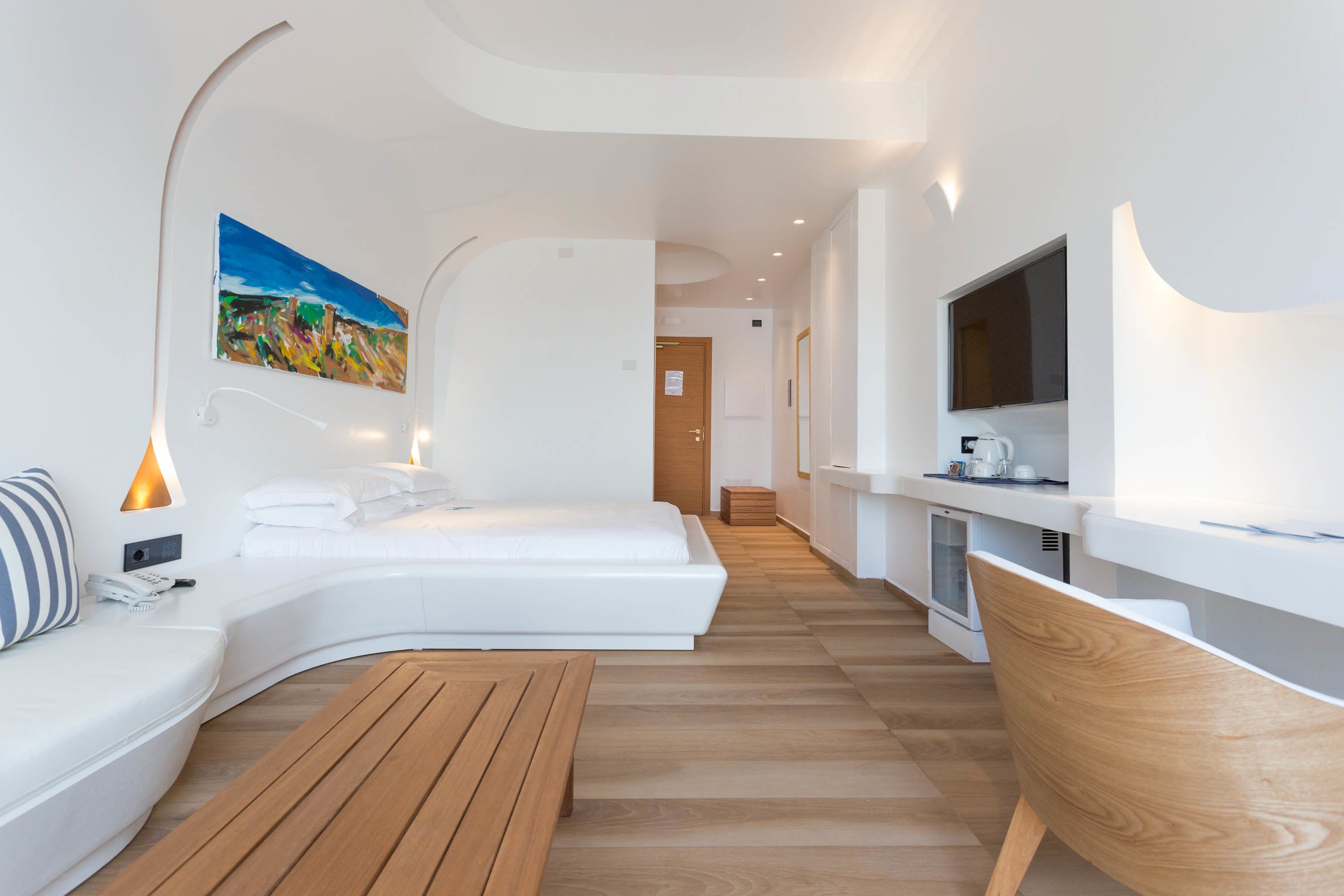 A modern hotel room with a curved bed, a wooden bench, a TV, and a painting on the wall.