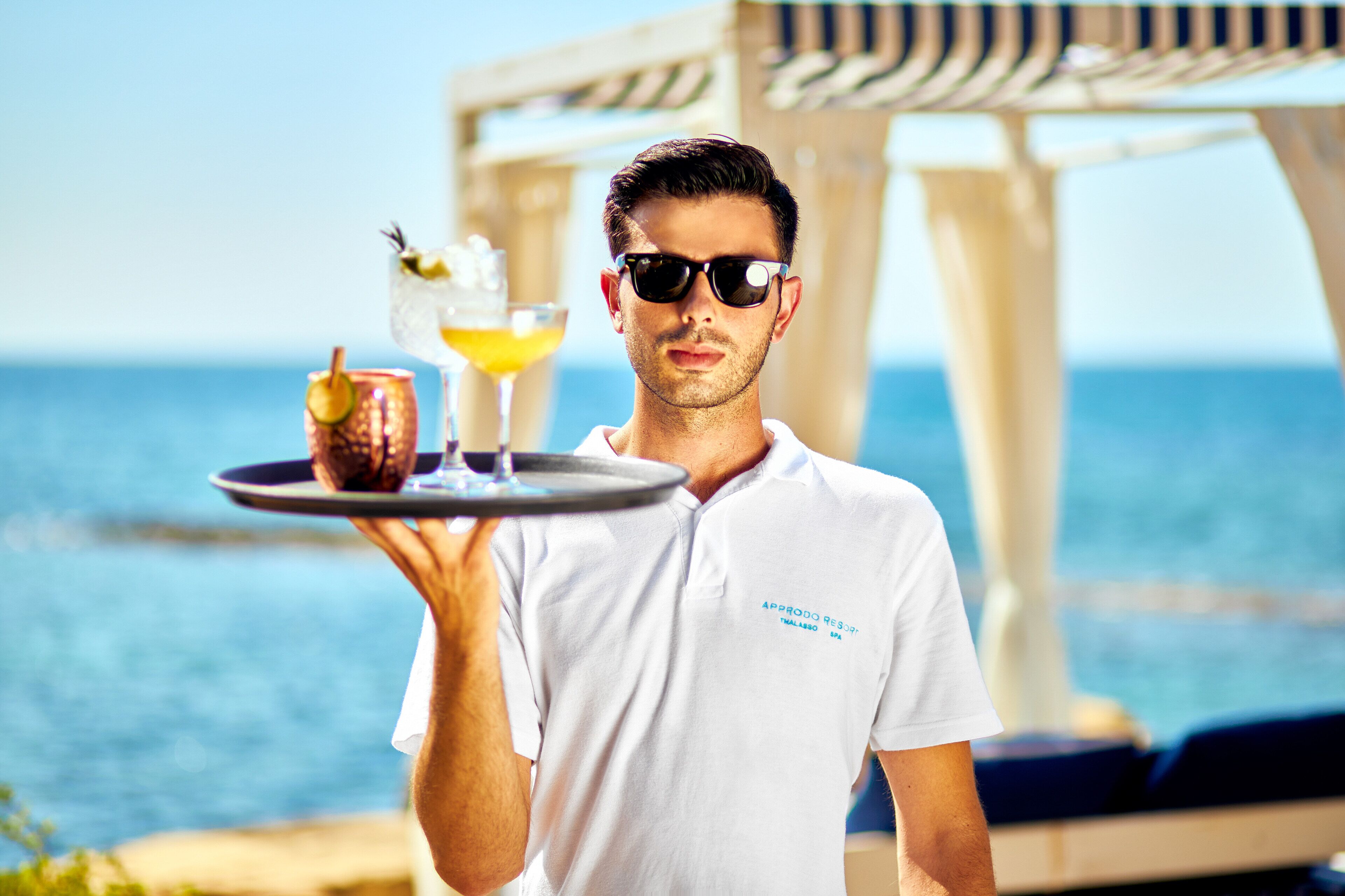 A person holding a tray with a drink and a dessert, standing near a pool with a view of the sea.