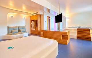 Una habitación de hotel moderna con una cama grande, un banco de madera, un televisor y un baño con bañera y ducha.