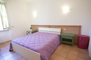 A bedroom with a bed, a nightstand, a green table, and a red trash bin. - Appartamento Mono Serenella (Portoferraio)